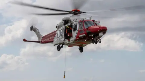 A red and white HM Coastguard helicopter is in the air. A rescuer holding a rescue rope out of the side of the helicopter.