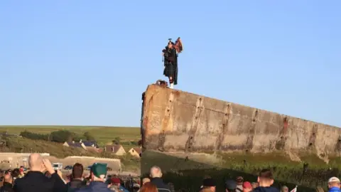 PA A lone piper plays on the Mulberry harbour at Arromanches in Normandy