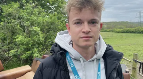 A young man with blond hair stands outside, looking into the camera lens. He is wearing a grey hoodie and navy coat, as well as a light blue lanyard which says "We're Kirklees" on it. Behind him is a pile of brown sofas, and a luscious green landscape in the background.