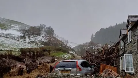 Emyr Owens Sebuah rumah batu sebagian dikaburkan oleh tumpukan pohon tumbang yang telah menabrak dinding luar. Perabotan kayu dapat terlihat tergeletak di lantai di luar rumah sementara Honda perak diparkir di luar. Bukit -bukit yang ditutupi dengan salju ringan dapat dilihat di latar belakang.