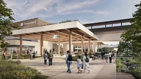 East West Rail A computer generated impression of what Tempsford station might look like. The image is framed on the left and right by greenery and there are a few dozen people walking into and out of a well-lit station entrance, which is under a broad canopy, held up by large square pillars. The railway symbol and the word "Tempsford" can be seen on the side of a building to the left of frame.