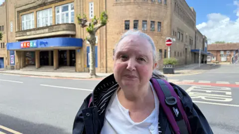 Woman with grey hair looking at the camera. She is a coat and has a backpack on. She is stood opposite the the Mecca Bingo Building. There's a keep clear bus stop sign in the road. It's a sunny day in the background but she is stood in the shade.