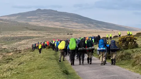 A large group of walkers set off with backpacks and camping gear. They are walking along a path on the moors on a grey day. 