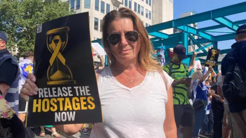 A posed photo of Julie at Sunday's protest. She is a middle-aged woman with mid-length light brown hair, wearing a v-neck white t-shirt and aviator sunglasses. She is smiling and holding a black and yellow sign that says 'release the hostages now'. Protesters are dotted around behind her.