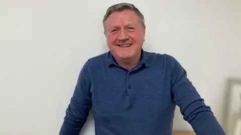 Kate Moser Andon/BBC Dennis Jones, wearing a blue long-sleeved polo shirt - smiles for the camera 