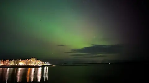 Curtis/BBC News NI Weather Watcher The sky lit up green with Northern Lights. There is a body of water in the bottom half of the image with a row of homes lit up to the left of the water. The reflection of the lights from the homes is visible in the water. 