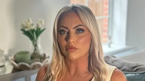 BBC A woman with long blonde hair and blue eyes is looking directly into the camera, unsmiling. She is wearing a light-coloured sleeveless v-neck top. The background is out of focus but she is sitting on a grey sofa with light green cushions, behind her is a table with white flowers on it and a window.
