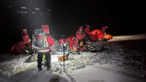 Assynt Mountain Rescue It is dark and rescue volunteers are in red suits on a sow covered mountain. There is snow blowing around them. There is an orange tent on the ground aglow with torch light. They have walking poles, head torches and large black backpacks.