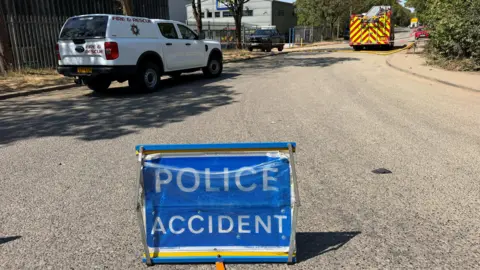 Richard Knights/BBC A police accident sign on a road, with a white fire van and a fire engine in the distance. A building is to the right. A yellow hose is running across the road. A metal fence is to the left and trees to the right. 