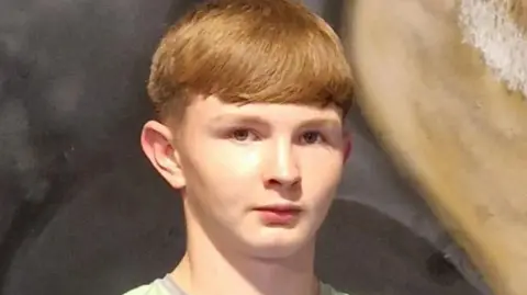 A male teenager with ginger hair looking at the camera. He's wearing a grey t-shirt with green panels on the arms.