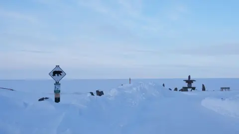 Eloise Alanna/BBC News An image showing the frozen, snow blanketed shore of the Hudson Bay as seen from Churchill. There is a sign with a polar bear symbol alerting locals. In the far right side of the photo, there is a stone inukshuk structure next to a bench. 