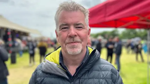 Mark Jones has short grey hair and a grey goatee, wearing a black waterproof jacket.