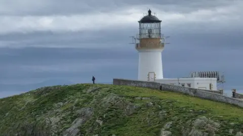 Chris Downer/Geograph The lighthouse is a stubby tower painted white with the glassed top and light behind it. The lighthouse is on a clifftop overlooking the sea. A figure is standing below the lighthouse.