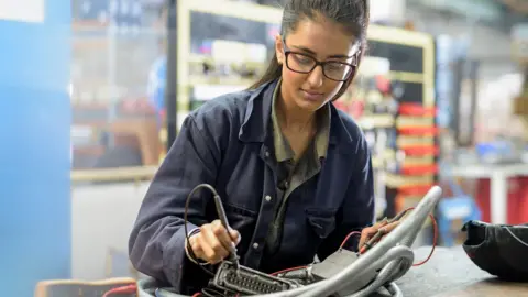 Getty Images Stock photo of an employee with long hair and glasses working on a soldering desk in an engineering environment while wearing work overalls.