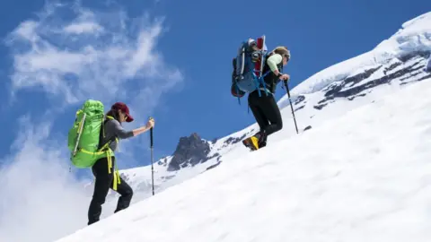 Cavan Images/Gabe Rogel via Getty Images Two women in outdoor gear with sticks and big rucksacks walk up a mountain in the snow