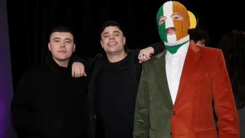 Getty Images The young men stand together. One is wearing a green, white and orange balaclava and suit. 