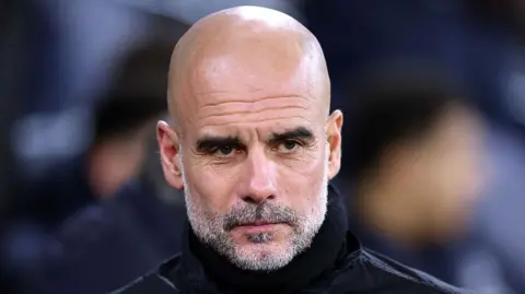 Pep Guardiola has a shaved head and grey beard, and wears a black jacket