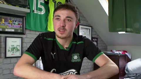 A man with short dark hair and slight facial hair sits in a room decorated with Plymouth Argyle football memorabilia. He is wearing a black and green team jersey. Behind him, framed pictures, football shirts, and a grey brick-patterned wall are visible.