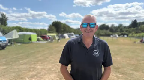 A man with short hair wearing black is smiling at the camera. A field behind him on the photo occupies much of the image, with vehicles blurred in the background parked by a hedge on the left.