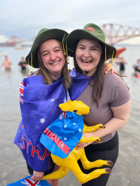Two women embrace while standing in the Firth of Forth - one has an Australian flag draped around her shoulders
