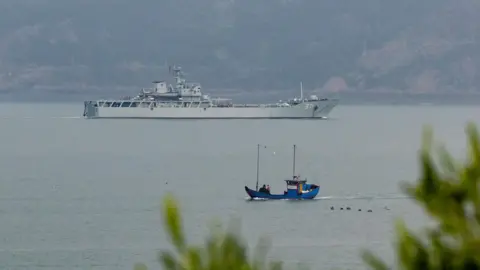 Reuters A white military ship sailing in the water. In the background are sloped lands, and in the foreground is a small blue boat.