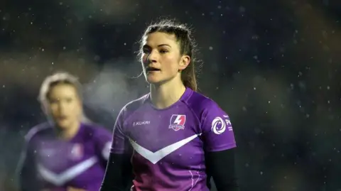 Helen Nelson playing for Loughborough Lighting in a purple rugby kit, with black long sleeves. Her hair is tied back and there is another player in the background. It is raining in the image.