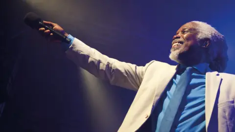 Dean Chalkley Billy Ocean on a stage during a performance. He is smiling and has his microphone pointed away from him.