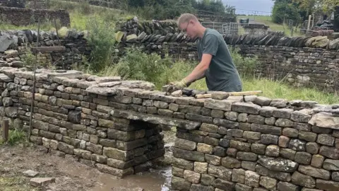 Phil Dolphin A young man wearing a green T-shirt, glasses and work gloves builds a dry stone wall enclosure in a grassy field.