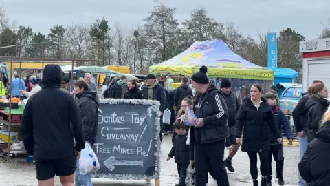 A sign says Santas Toy giveaway and free mince pie. Stalls can be seen in the background and people are milling around wrapped up in winter coats.