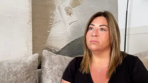Rachel Bertenshaw is sitting on a grey sofa. She is wearing a black top and has long blonde-brown hair. 