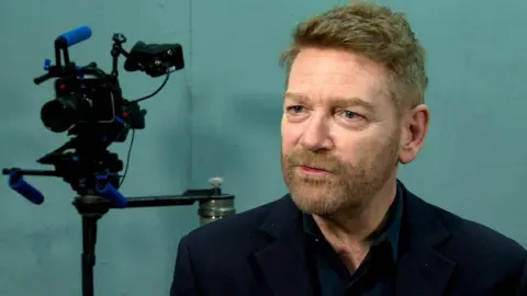 Sir Kenneth Branagh wearing a black shirt open at the collar. He has a short light-brown full beard and short hair.