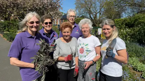 A group of six women, three of whom are wearing purple polo shirts and several of whom are holding plants or gardening implements. They are looking towards the camera and smiling for the photograph.
