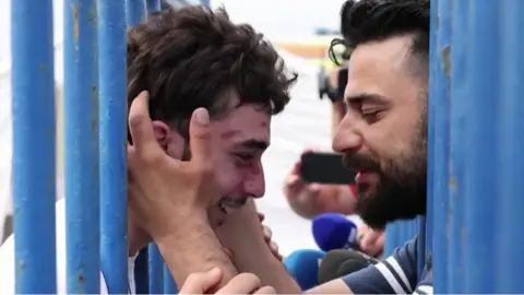 Two men smiling and embrace, with two rows of fences separating them