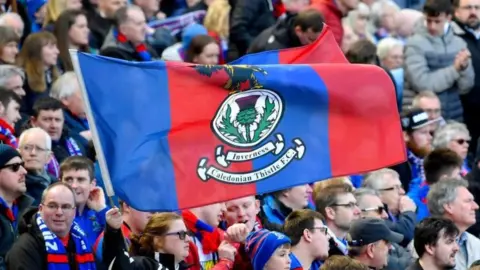 SNS Large bluish  and reddish  caley thistle flag, with thistle emblem successful  the centre, is held by a instrumentality   among a assemblage  of supporters astatine  a match.