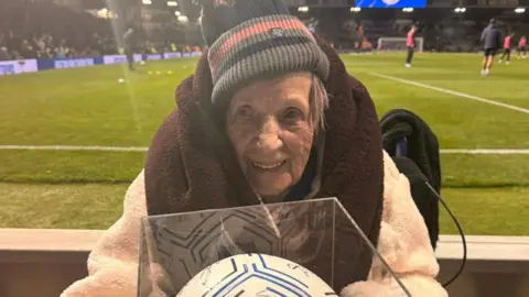 Joan, an elderly woman wearing a woolly hat and with a blanket wrapped around her neck, smiles in the direction of the camera. She is sat in front of a football pitch, where a few players can be seen dotted around. 