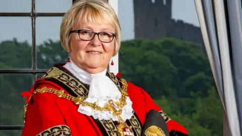 Official portrait of a woman with short blonde hair and glasses, wearing gold mayoral chains and red robes with black and gold detail.