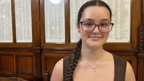 Savannah Shellard smiles at the camera indoors. She is wearing glasses and has a long plait, with wood panelling and framed rolls behind her.