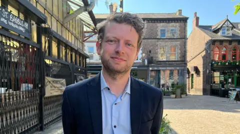 MP Connor Rand in Altrincham town centre. Altrincham Market's black gated fence is next to him and there are other town centre buildings in the background.