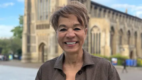 Cameron Noble/BBC Tessa McWatt standing outside The Forum in Norwich in front of a church which is out of focus. She is looking directly at the camera and is smiling. She has short brown hair.