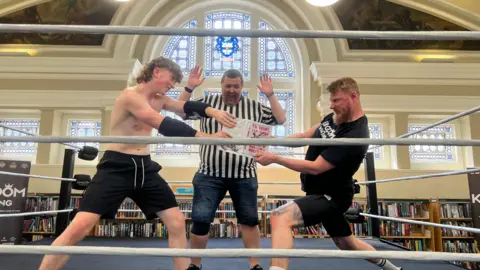 Jessica Bayley/BBC A boy in black shorts and no shirt stands on the left, a man wearing black shorts and a black T-shirt is on the right. They are both holding onto a book, fighting over it. Behind them stands a ref. All three are stood in a boxing ring. Behind them is a stained glass window and bookshelves.