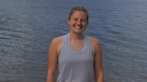 Freya Shepherd Freya is standing in the middle of the shot and is  smiling broadly. She is wearing a grey, sleeveless top and black shorts and Loch Ness is behind her. 
