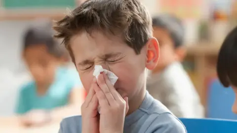 Getty Images Child sitting indoors with hands near mouth, wearing a light blue top, in a classroom setting with other children and colorful background elements.