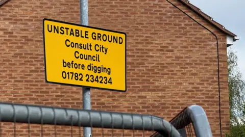 A yellow sign with black writing on it indicating the ground is unstable. In the distance is a brick building.