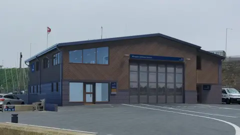 A generated image of what the new site is expected to look like. It is a two-storey building with dark wooden cladding on the outside and large glass windows at the front.