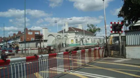 The level crossing, protected by red and white gates, at Portslade station, which has two platforms and a white and green building