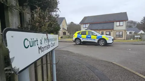 A police car parked in Morrison Court. The SUV is white white blue and yellow markings. It is parked outside a semi-detached house. There is a sign in the foreground that says "Morrison Court" in English and Gaelic.
