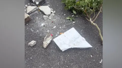 Debris including broken concrete, a white marble tile and a leafy green plant scattered on asphalt