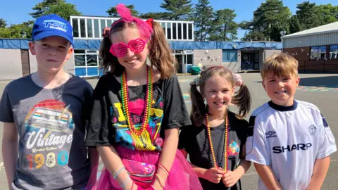 Four children in brightly coloured clothing stand in a school playground and smile at the camera