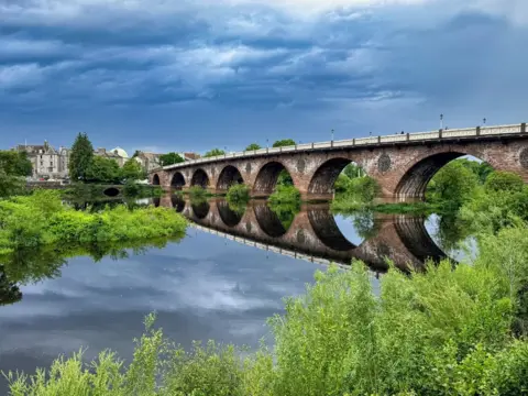 Brian Johnston A stone bridge spanning over the River Tay and reflected in the water. There are green bushes and trees along the banks of the river.
