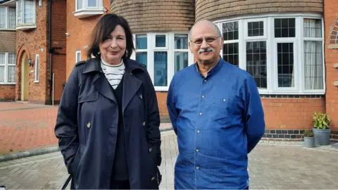 Birmingham City Council A woman and a man standing on a driveway in front two residential properties. The woman is in a navy trench coat and the man is in a denim shirt. They are both smiling into the camera. 